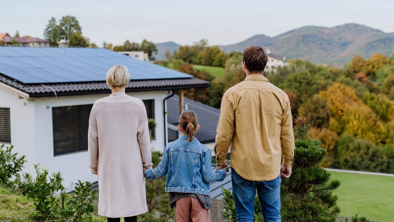 Are solar panels worth it? A family of three standing hand-in-hand with their backs turned to the camera looking at solar panels on the roof of a house with mountains in the background.